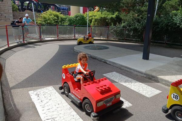 A little girl drives a red Lego car at Legoland Germany’s driving school, looking focused on the road ahead. Other kids in Lego cars are also cruising around the track, while parents watch from the sidelines. It’s a fun, mini driving adventure made just for kids!