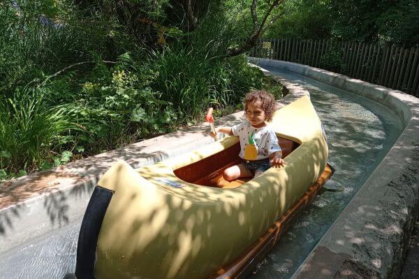 A little girl enjoys a canoe ride at Legoland Germany, holding a small paddle and smiling. The canoe floats along a winding water channel surrounded by green plants and trees. Sunlight filters through the leaves, making it a perfect day for adventure.