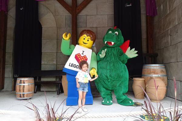 A young girl poses with two life-sized characters at Legoland Germany—a friendly Lego figure wearing an "I ❤️ LEGO" shirt and a cheerful green dragon with red accents. The girl holds hands with the Lego figure while the dragon waves. The background features a medieval-style setting with wooden barrels and stone walls.