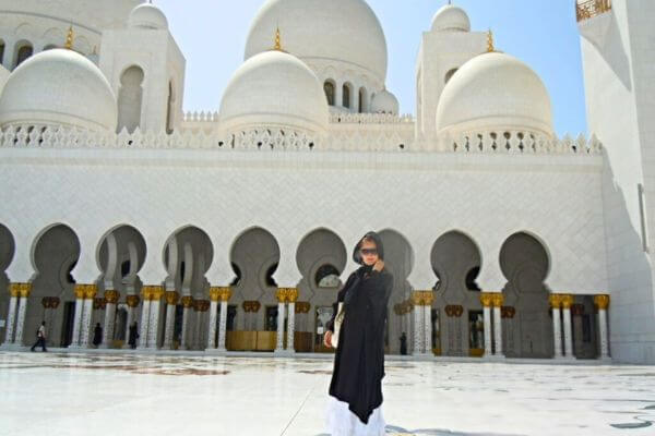 A woman standing in Sheikh Zayed Grand Mosque, Abu Dhabi A woman in black and white attire (traditional Arabic clothes with hijab) stands in the Sheikh Zayed Grand Mosque in Abu Dhabi under a clear blue sky.