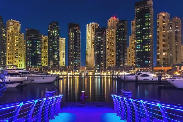 Nighttime cityscape of illuminated skyscrapers reflecting in Dubai Marina. Luxury yachts are docked, and a blue-lit pathway leads to the water.