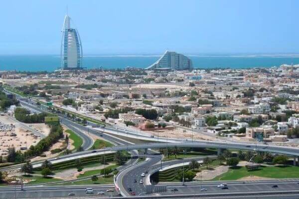 Aerial view of Dubai with a modern highway and interchanges in the foreground. In the background, Burj Al Arab Hotel and ocean horizon under a clear blue sky.