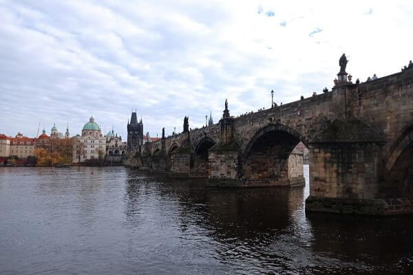 Charles Bridge in Prague stretches across the Vltava River under a cloudy sky, with people walking along its length and statues lining the sides. Historic buildings, including a dome and a tower, are visible in the background on the left.