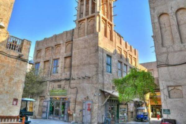 Al Bastakiya Dubai A quiet street in Al Bastakiya, Dubai, showing traditional sand-colored buildings with wooden wind towers and small shops below. The scene is calm with a few trees and signs inviting visitors to explore.