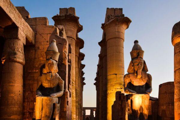Large pharaoh statues and stone columns at Luxor Temple in Egypt, lit up at sunset.