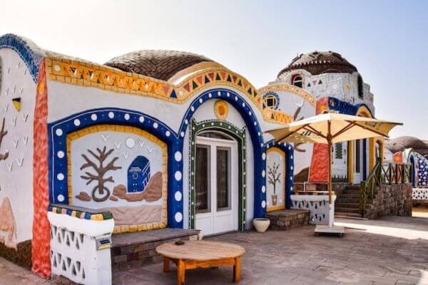 Nubian Village in Aswan, Egypt Nubian Village in Aswan, Egypt. It shows a colorful, dome-shaped building with detailed patterns and vibrant designs, including painted trees and geometric shapes. A small wooden table and a large umbrella stand in the foreground on a stone patio, creating a cozy outdoor space.