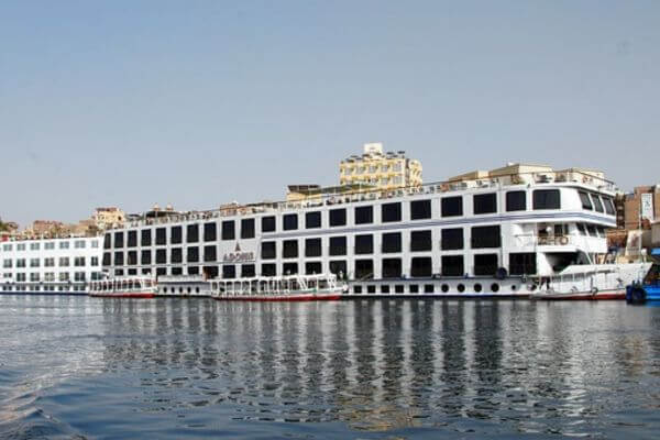 Nile River Cruise, Egypt A large white river cruise ship docked along the River Nile, with small boats tied beside it. Buildings and a clear blue sky can be seen in the background.