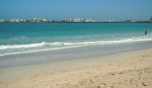 Umm Suqeim Beach in Dubai A peaceful view of Umm Suqeim Beach in Dubai, with gentle waves rolling onto the sandy shore. The calm blue water stretches out to a line of buildings and a long bridge in the distance.