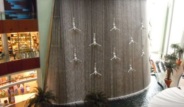 Human Waterfall in Dubai Mall A large indoor waterfall at Dubai Mall with silver human-shaped sculptures diving headfirst in streams of falling water. The wall curves into a pool surrounded by palm trees and nearby shops.