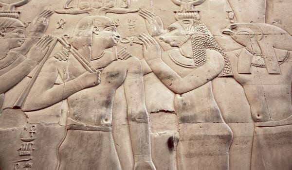 Temple of Kom Ombo, carvings on the wall Close-up of an ancient Egyptian wall carving showing figures in profile, including a deity with a crocodile head and another with a falcon head, interacting with human figures. The scene is carved into stone with detailed outlines and symbols.
