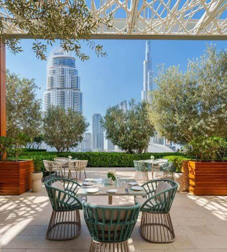 A restaurant at Address Fountain View Outdoor dining area at a restaurant in Address Dubai Mall Hotel with a view of the Burj Khalifa and nearby towers framed by greenery.