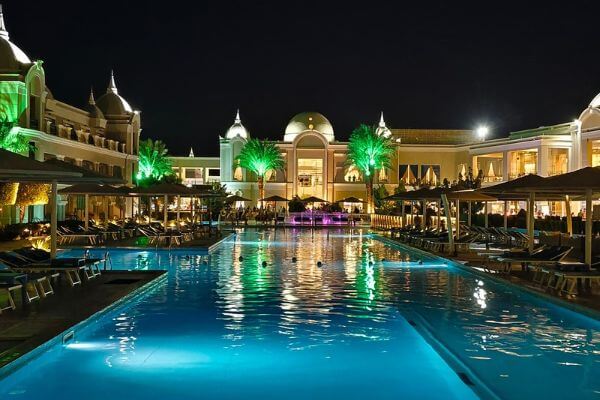 Beach resort in Hurghada, Egypt Beach resort in Hurghada, Egypt, at night, with a large pool lit up and palm trees glowing green in front of the main building.