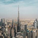 Aerial view of Dubai with Burj Khalifa at the center. Surrounding are numerous high-rise buildings under a partly cloudy sky.