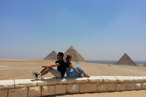 Near Pyramids of Giza Couple sitting back to back on a stone wall near the Pyramids of Giza in Egypt, with the pyramids behind them under a clear sky.