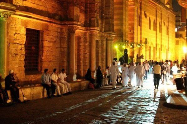 This is a lively street in Cairo at night, with warm lights illuminating the historic stone buildings. People are sitting along the wall, while others walk and gather in small groups, dressed in traditional and casual clothing. The cobblestone street reflects the glow of streetlights, adding to the vibrant atmosphere.
