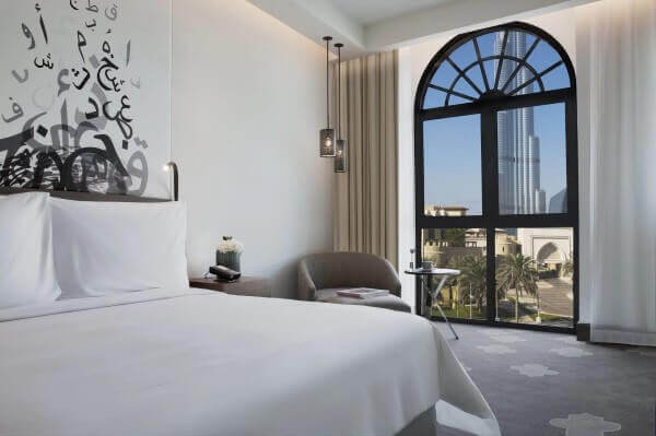 The Heritage Hotel, Autograph Collection, Burj view rooms Modern hotel room in the Heritage Hotel, Autograph Collection, Dubai with a white bed, abstract Arabic calligraphy on the wall, large arched window revealing a cityscape with a Burj Khalifa.