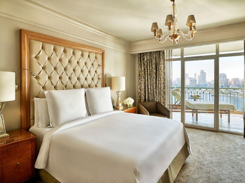 Suite bedroom at the Four Seasons Hotel Cairo at Nile Plaza, with a king bed, a Nile view balcony, and city skyline in the background.