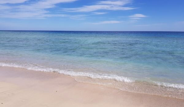 Abu Dabbab Beach, Egypt Tranquil Abu Dabbab Beach with clear turquoise waters gently lapping against a sandy shore under a bright blue sky with scattered white clouds.