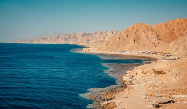 Blue Hole, Dahab A calm view of Blue Lagoon beach in Egypt with clear blue water meeting a sandy shoreline. Rocky hills stretch along the coast under a bright blue sky.