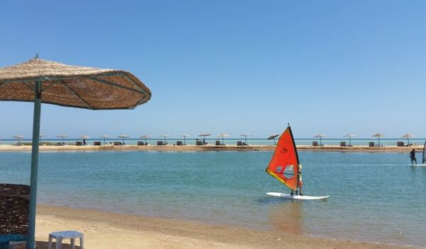 El Gouna, Egypt A person sails a small board with a bright orange sail in the calm waters of a beach in El Gouna near Hurghada, Egypt. Straw umbrellas line the sandy shore in the background under a clear blue sky.