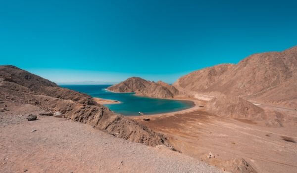 Fjord Bay, Egypt Fjord Bay in Egypt shows calm blue water surrounded by rugged brown mountains under a clear sky. A small sandy strip curves along the edge of the bay, creating a peaceful spot between the hills.