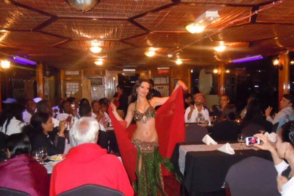 A Belly dancer on Dhow cruise in Dubai A belly dancer performs on a Dhow cruise in Dubai, moving gracefully with a red veil. Guests sit around dining tables, watching the show and enjoying the lively atmosphere. The warm lighting and smiles add to the fun evening vibe.