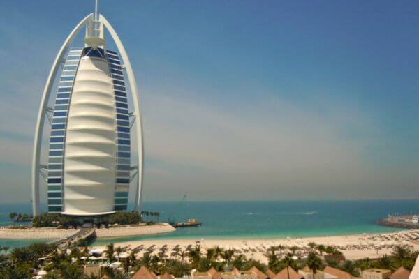 Burj Al Arab, Dubai The Burj Al Arab hotel stands by the sea in Dubai, shaped like a sail and surrounded by clear blue water. The sandy beach and palm trees add a calm and sunny vibe to the scene.