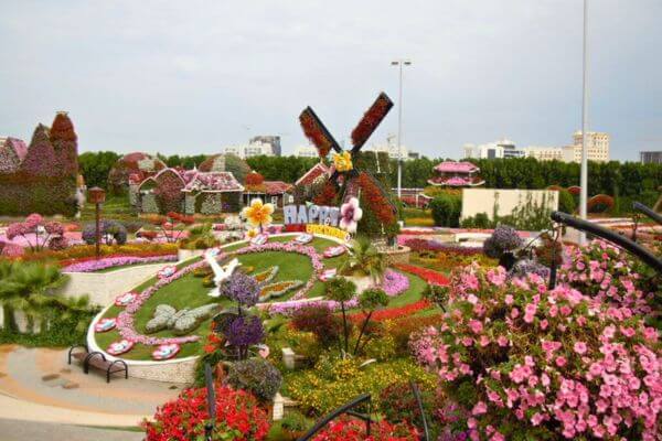 Dubai Miracle Garden Colorful flower displays fill the Dubai Miracle Garden, with a windmill and the words “Happy Birthday” made of blooms at the center. The garden paths wind through bright flower beds, shaped plants, and small structures covered in blossoms under a clear sky.
