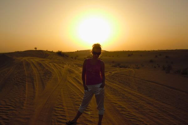 Dubai desert safari at sunset A person stands on sandy dunes during a desert safari in Dubai, with the sun setting low behind them. The sky glows warm and golden, and faint tire tracks mark the soft sand around them.