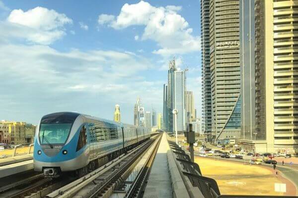 Dubai Metro A sleek Dubai Metro train moves along elevated tracks surrounded by tall city buildings. The sky is bright with a few clouds, and cars can be seen on the road below.