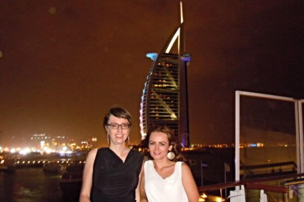 Rooftop bar, Dubai Two people smile on a rooftop bar in Dubai at night, with the Burj Al Arab lit up in the background. City lights and reflections from the water add to the lively atmosphere.