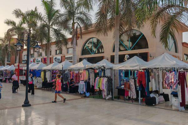 Outdoor clothing stalls set up in front of shops in New Cairo, Egypt, with palm trees and people walking by.