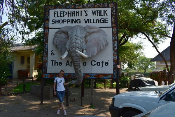 Me in Victoria Falls Town in front of a sign for Elephant's Walk Shopping Village
