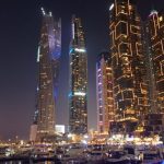 Dubai Marina at night with tall, brightly lit skyscrapers reflecting on the water. Boats and yachts are docked along the marina, adding to the lively evening atmosphere.