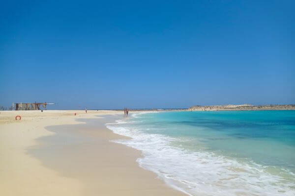 Almaza Bay Beach, Marsa Matrouh, Egypt Golden sand meets clear blue water at Almaza Bay Beach near Marsa Matrouh, Egypt, with gentle waves rolling to shore under a bright blue sky. A small beach hut and a few people can be seen in the distance enjoying the peaceful shoreline.