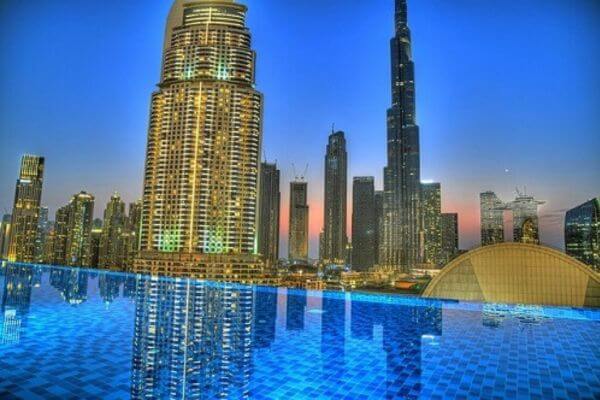 Skyline of Dubai at dusk with illuminated skyscrapers, including Burj Khalifa. A reflective infinity pool in the foreground adds elegance.