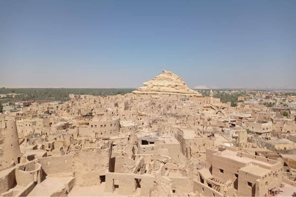 A view from the top of Shali Fortress in Siwa View of the old mudbrick buildings of Shali Fortress in Siwa, Egypt, with the white rock hill of Gebel al-Mawta rising in the background under a clear blue sky. The photo shows the dense layout of the ancient town surrounded by desert and greenery in the distance.
