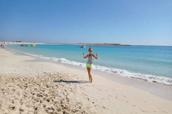 Almaza Bay beach A woman poses playfully on the soft sandy shore of Almaza Bay in Marsa Matrouh, Egypt, with clear turquoise water gently lapping at her feet. The beach stretches into the distance under a bright blue sky, with a few boats and beachgoers visible in the background.