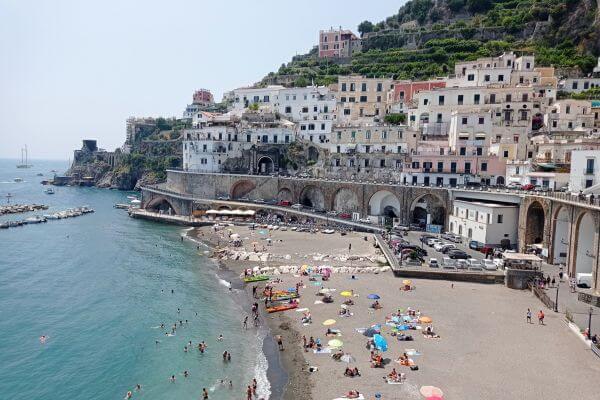 Atrani Town, Amalfi Coast, Italy A sandy beach in Atrani, Italy, with colorful umbrellas and people swimming in the clear water. Behind the beach, white and pastel houses rise up the hillside along the Amalfi Coast.