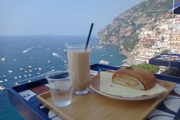 Breakfast with a view in Positano, Amalfi Coast A wooden tray holds a pastry, a glass of iced coffee, and a cup of water on a balcony table. Behind it is a clear view of the blue sea filled with boats and the hillside town of Positano along the Amalfi Coast.