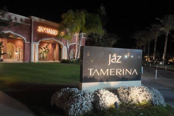 Jaz Tamerina Hotel A nighttime view of the Jaz Tamerina Hotel entrance in Almaza Bay, Marsa Matrouh, Egypt. The hotel sign is lit up in front of a well-kept lawn with palm trees and a warmly lit building in the background.