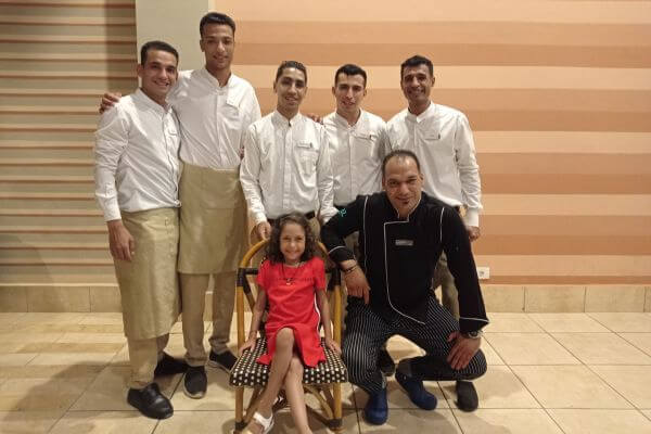 Jaz Tamerina, restaurant's staff A group photo of the restaurant staff at Jaz Tamerina posing with our daughter, who is smiling while sitting on a chair in front. Everyone looks happy and relaxed, standing against a striped wall indoors.