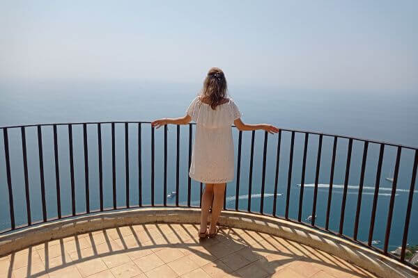 Ravello Cimbrone Garden A woman in a white dress stands at the railing of the infinity terrace in Villa Cimbrone Garden, in Ravello, looking out at the wide blue sea. Boats can be seen leaving white trails across the water below.