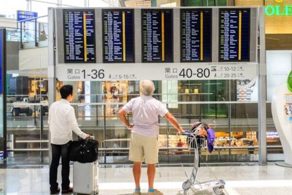 Traveling, airport Two travelers stand in front of a large flight information display board at an airport, scanning the screen for departure details. One man has a suitcase in hand, while the other leans on a luggage cart piled with bags and a neck pillow.
