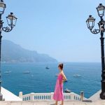 A woman in a pink dress stands between two black lampposts at a viewpoint in Atrani, looking out at the sea with boats and distant mountains on the Amalfi Coast