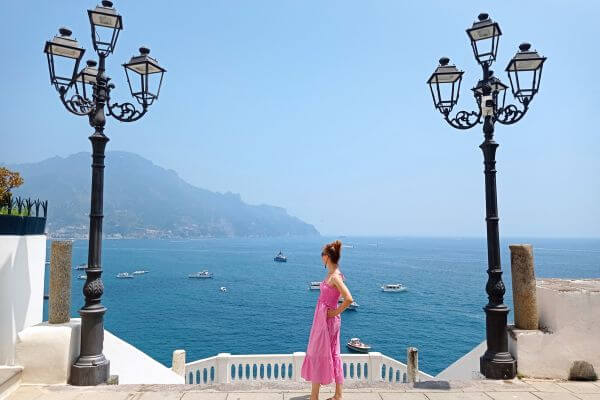 Atrani Town in Amalfi Coast A woman in a pink dress stands between two black lampposts at a viewpoint in Atrani, looking out at the sea with boats and distant mountains on the Amalfi Coast.