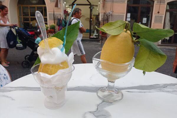 Lemon Sorbet in Lemon A cup of lemon sorbet served inside a hollowed lemon sits on a table next to a fresh lemon with green leaves in a glass dish. The background shows people walking by on a street in Amalfi, Italy.