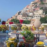 Colorful ceramic pots and tables decorated with lemon designs sit on a sunny terrace in Positano. Behind them, pastel houses climb the hillside overlooking the blue sea on the Amalfi Coast.