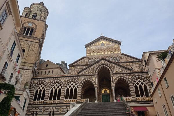 St. Andrea Cathedral, Amalfi Town, Italy The front view of St. Andrea Cathedral in Amalfi Town shows wide stone steps leading up to its striped arches and tall bell tower. The church stands between narrow buildings with a few people walking near the entrance.