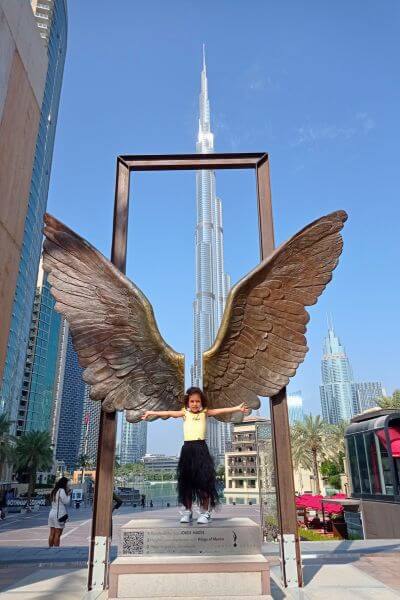 Wings of Mexico, Dubai A child stands smiling with arms outstretched in front of the bronze Wings of Mexico sculpture in Dubai, with the Burj Khalifa rising in the background. The sunny sky and modern city buildings create a bright, cheerful scene.